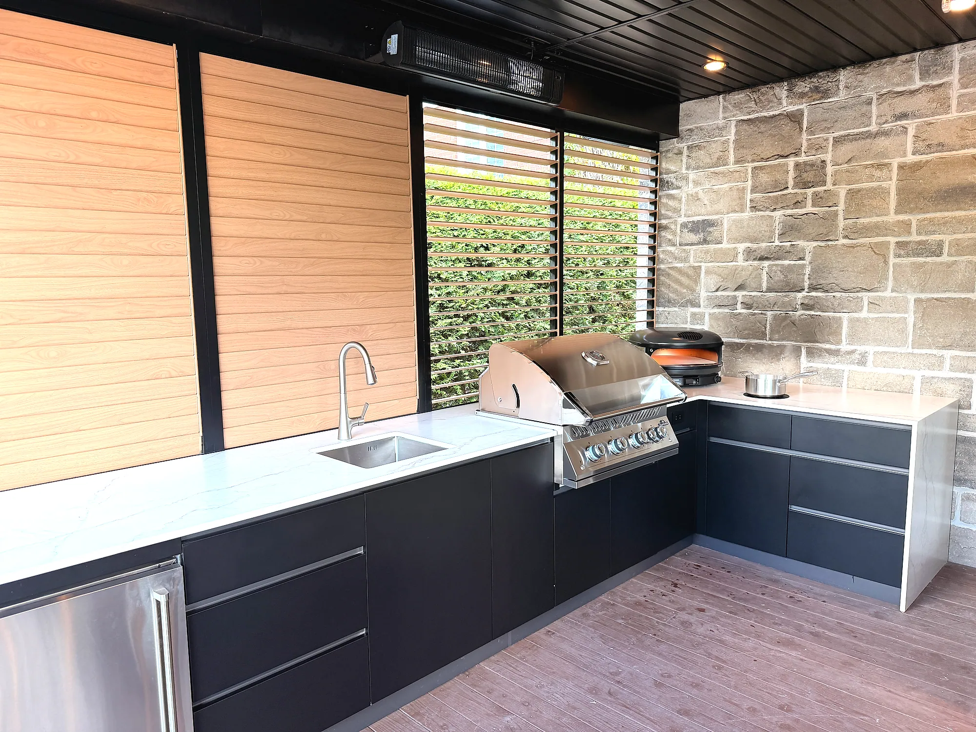 Cuisine extérieure en L sous terrasse couverte à Saint-Augustin-de-Desmaures, avec BBQ Napoléon, four à pizza Gozney, comptoirs Orastone Chalk et armoires Noir basalte, projet Mercier